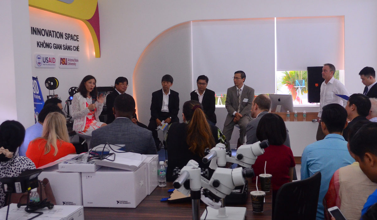 Photo of a group of people listening to a speaker in the corner with a caption of "Jeffrey Goss led a panel to discuss the developing next generation of innovators at the launch. Panelists included Do Van Dung, president of Ho Chi Minh University of Technology and Education, Sherry Boger, vice president of Intel Products Vietnam, Francis Tuan Anh Nguyen, evangelist at Microsoft, Nhan Nguyen, general director of FabLab Saigon, and Ban Hoang The, expert of technology transfer and innovation at Saigon Hi-Tech Park. Photographer: Quynh Giao"