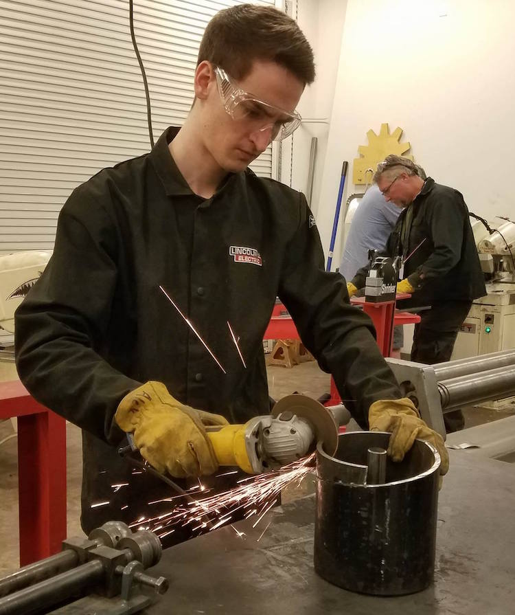 A photo of a student using a saw creating sparks with a caption of "Evren Uner preparing the auger press to create the spirals for the auger. Photo courtesy of Next Level Devils"