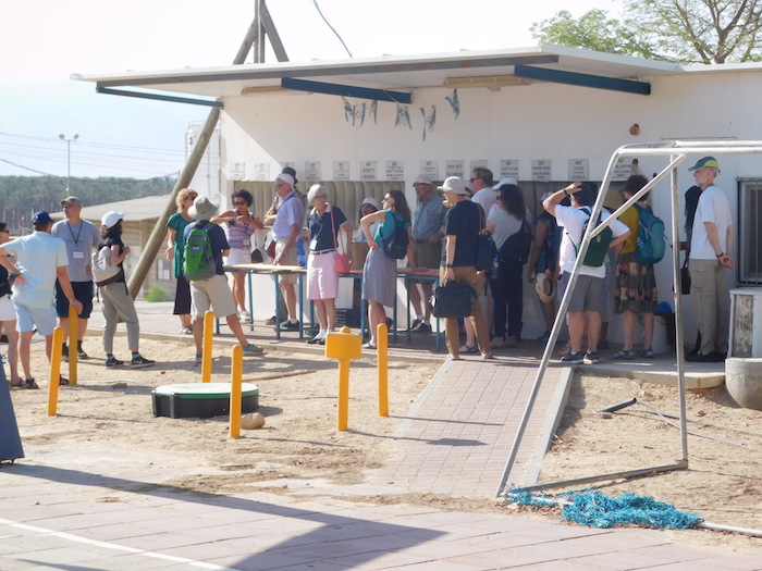 Photo of people standing outside a building. Caption: Tours of some of Israel’s kibbutzim, rural communal settlements, have been on the itineraries of the Faculty Fellowship trips, which seek to give visitors from the U.S. perspectives on daily life in Israeli society. The photo shows a tour group at the Kibbutz Ketura. Photographer: René Reinhard/Jewish National Fund.  