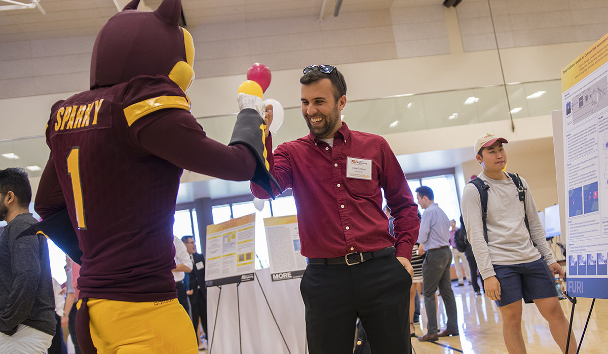 Photo of Sparky the Sun Devil giving a high five to a student. Caption: Cesar Tamayo explained to Sparky his research for an autonomous driving neural network algorithm that uses multiple cameras to help determine if a car is going to crash. Tamayo says he has learned so much about teamwork as part of his FURI research project, and has now submitted his second paper as an undergraduate student. Photographer: Marco-Alexis Chaira/ASU