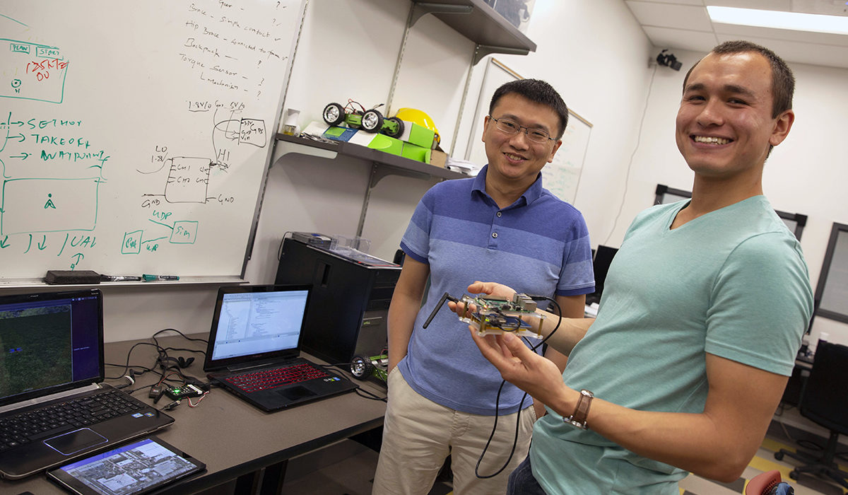 Photo of professor and student standing. Caption: YiZhuang “JJ” Garrard (right) poses with his mentor, Assistant Professor Wenlong Zhang in the lab. Garrard’s research focused on using low-cost and easy-to-use quadcopters for performing topological surveys. Garrard’s research was funded by the Kern Family Foundation for his entrepreneurial approach to his project. Photographer: Jessica Hochreiter/ASU