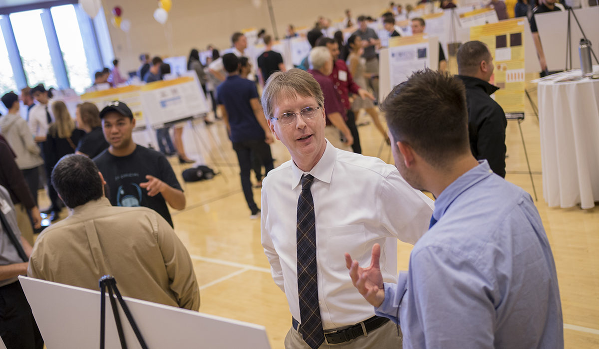 Photo of Dean Kyle Squires talking with a student. Caption: Fulton Schools Dean Kyle Squires attended the FURI Symposium to talk with students about their research. Squires says research enhances students’ engineering and technology education through developing important skills for their careers or graduate school: the ability to innovate, think independently, solve problems and defend their findings. Photographer: Marco-Alexis Chaira/ASU