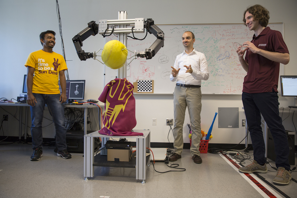 Three men with a robot that is holding a large plush tennis ball